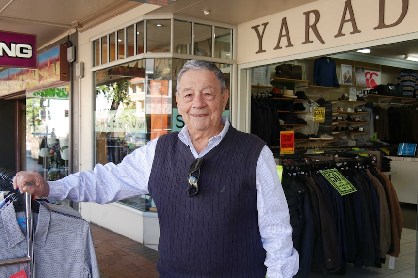 An older man stands outside a clothing store, smiling.