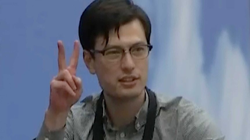 Alek Sigley holds up two fingers in the V for victory sign as he walks through an airport terminal.