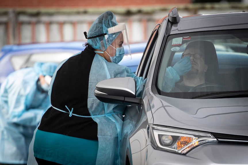 A woman wearing protective clothing tests a man in the drivers seat of a car for COVID-19.