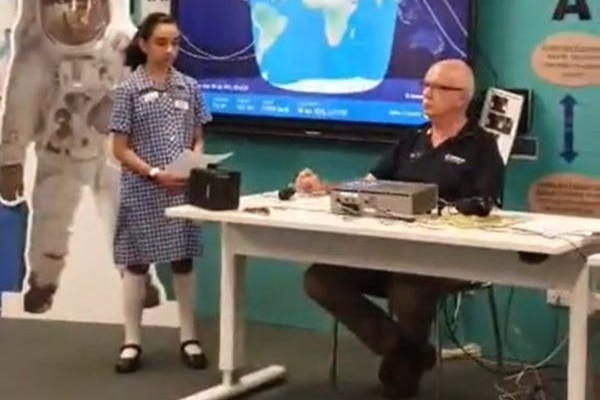 A girl wearing a school dress stands at a table with a man and a cut-out of an astronaut and a screen showing the earth