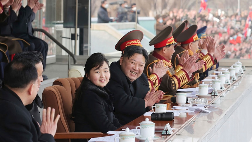 A beaming, heavy Korean man claps as he sits alongside a number of generals and a young, smiling Korean girl.
