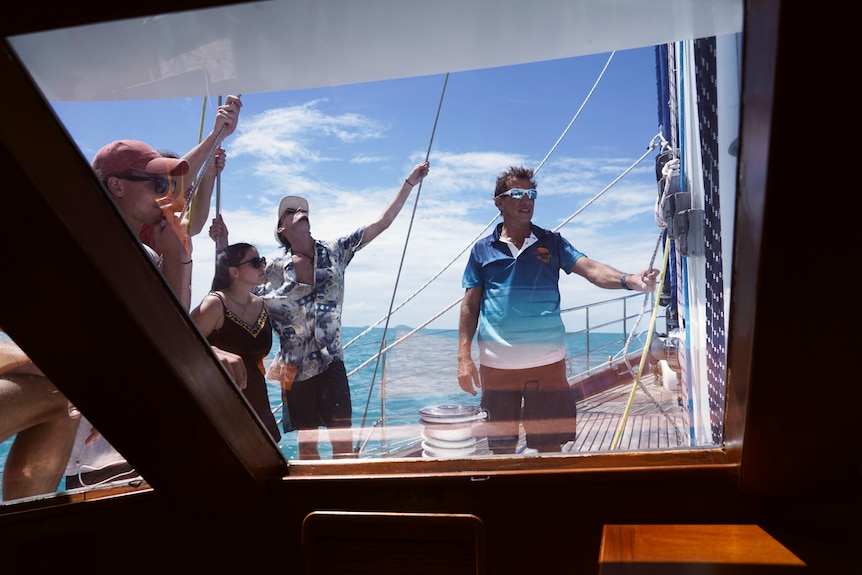 A man in a blue and white polo shirt stands on deck teaching his crew how to put the sail up. 