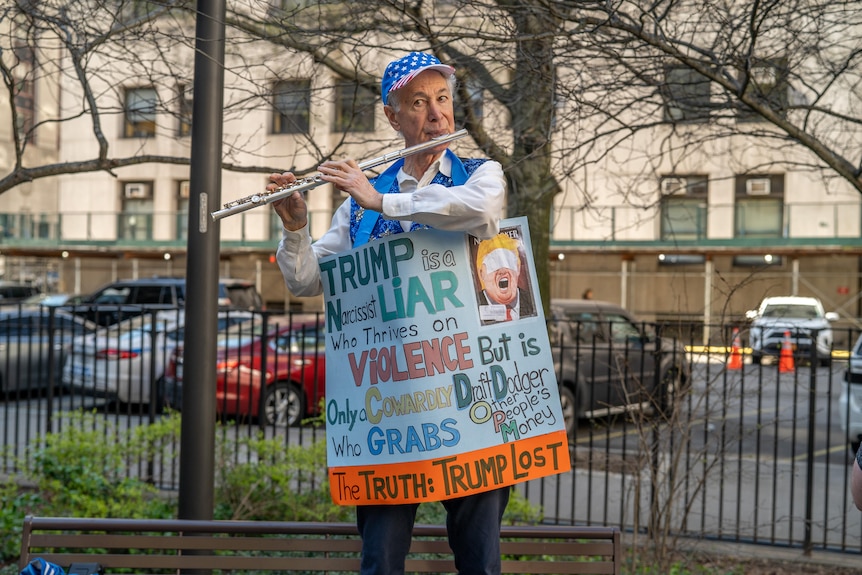 A demostrator plays a flute and wears a sign with anti-Trump slogans outside a courthouse.