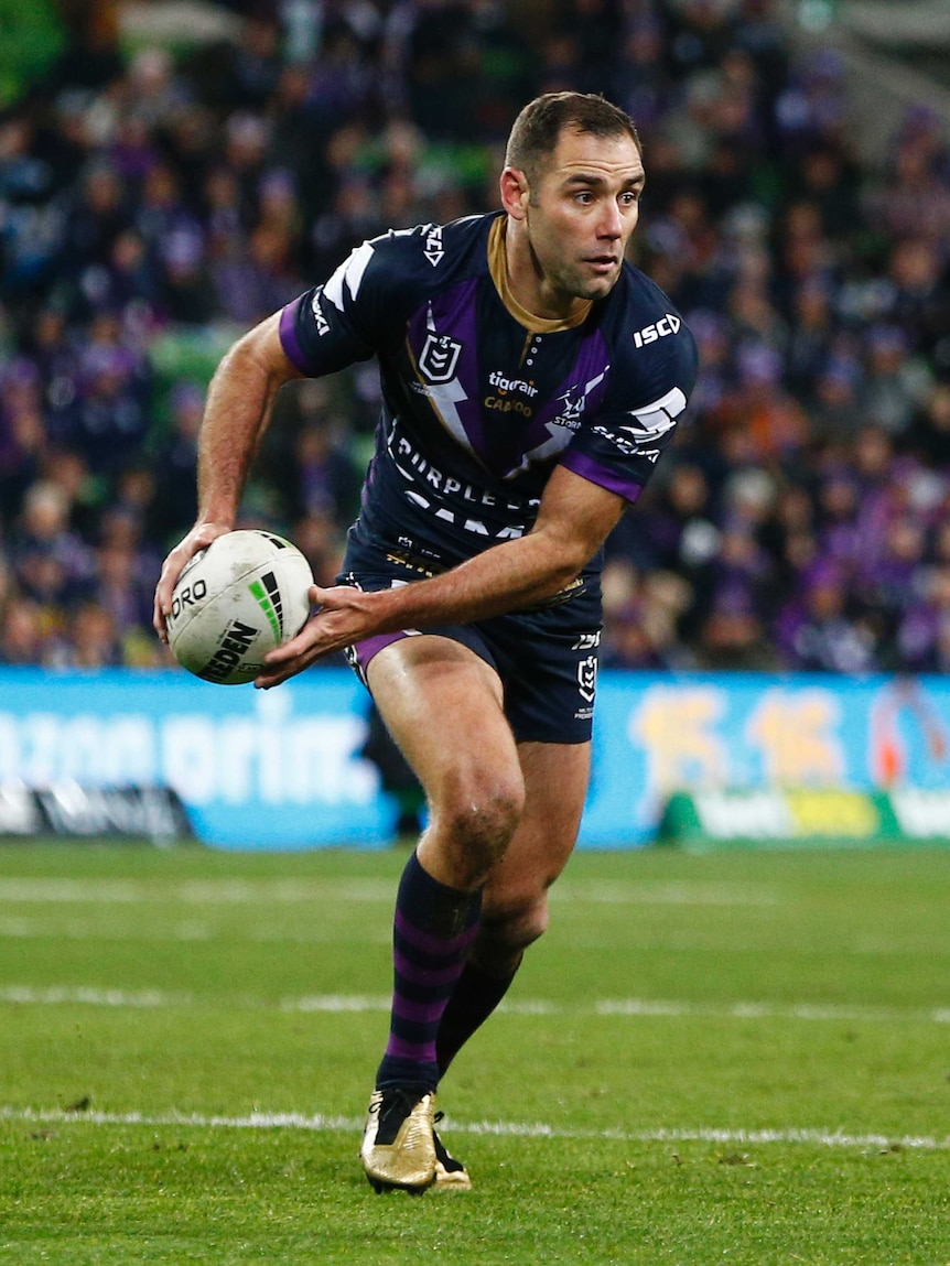 A male NRL player moves to his left as he holds the ball in both hands at waist level.