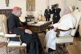 Priest in black robes and red cap sits opposite priest in white robes and white cap