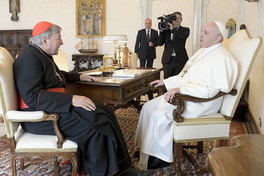 Priest in black robes and red cap sits opposite priest in white robes and white cap
