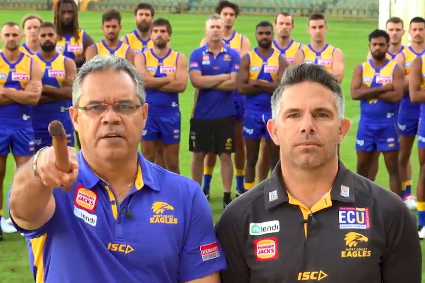 Two men in Eagles shirts, one pointing a stick at the camera, stand in front of players on a football oval.