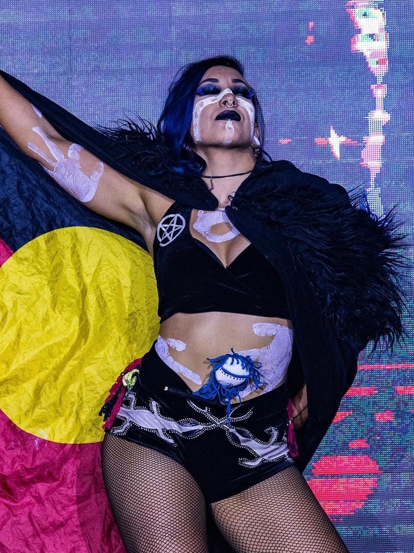 woman covered in body paint stands holding an Aboriginal flag in front of large tv screen.