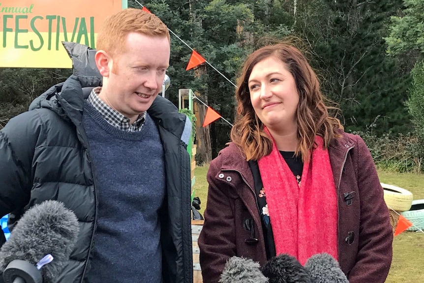 A man and a woman wearing heavy coats talk in front of several microphones outside with trees in behind