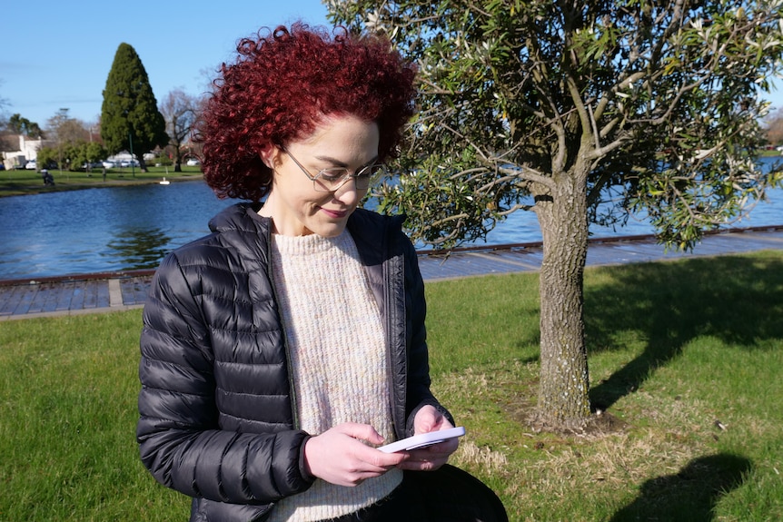 A woman with bright, curly hair sits near a waterway on a stunning day, looking at her phone.