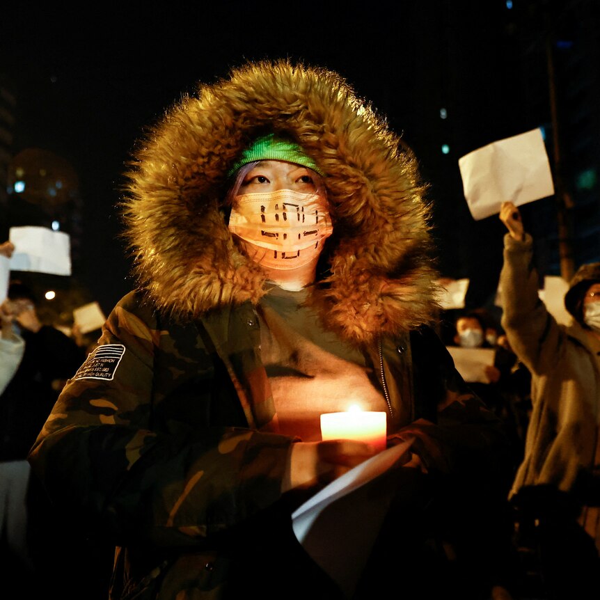A person holds a candle, as people gather for a vigil and hold white sheets of paper in protest.