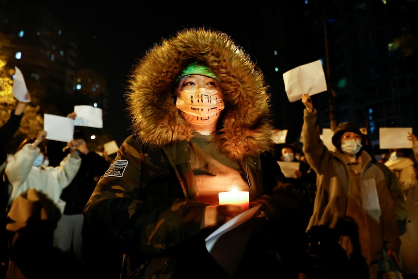 A person holds a candle, as people gather for a vigil and hold white sheets of paper in protest.