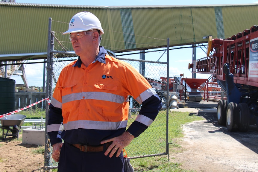 A man in a high-vis uniform shirt and hard hat stands with his hands on his hips, infrastructure behind him