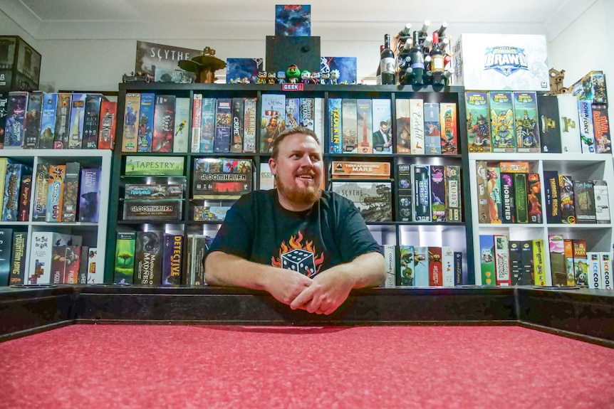 A man leans on a large table. Behind him are shelves of board games.