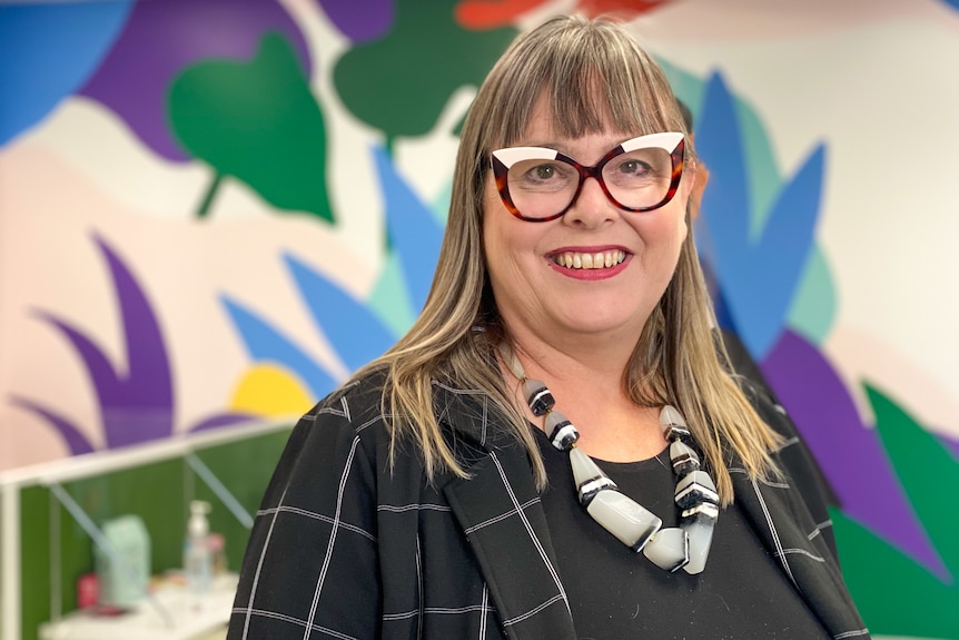 a woman with glasses is smiling at the camera. She wears a blazer and large necklace.