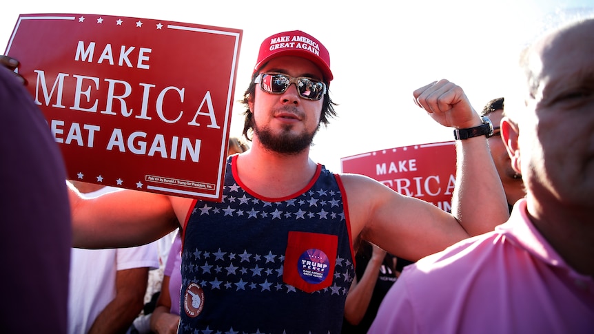 A man holds up a sign reading "make american great again" and flexes his muscles