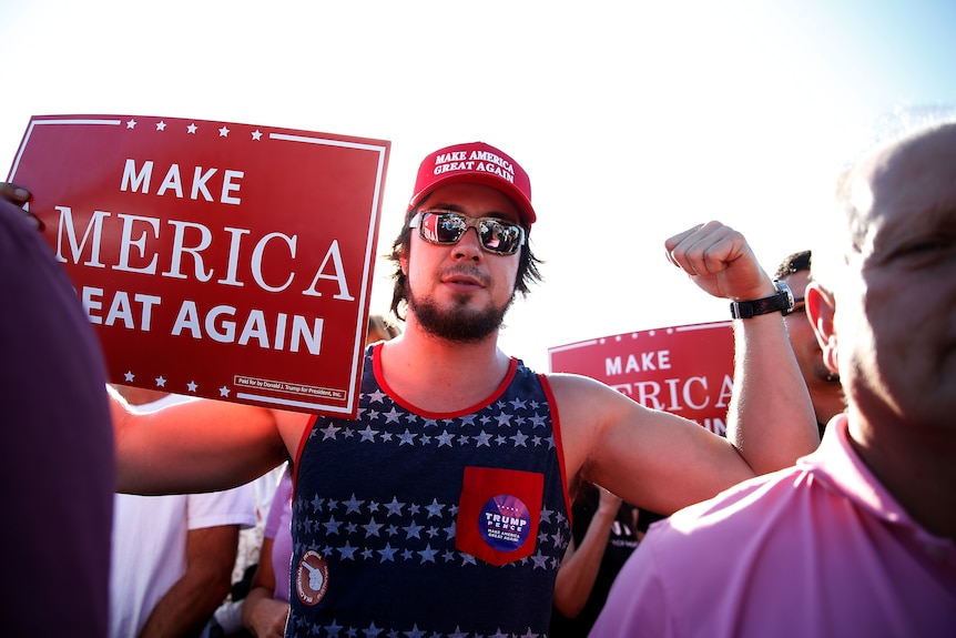A man holds up a sign reading "make american great again" and flexes his muscles