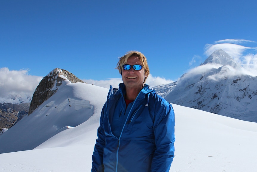 John All stands on a mountain surrounded by snowy peaks.