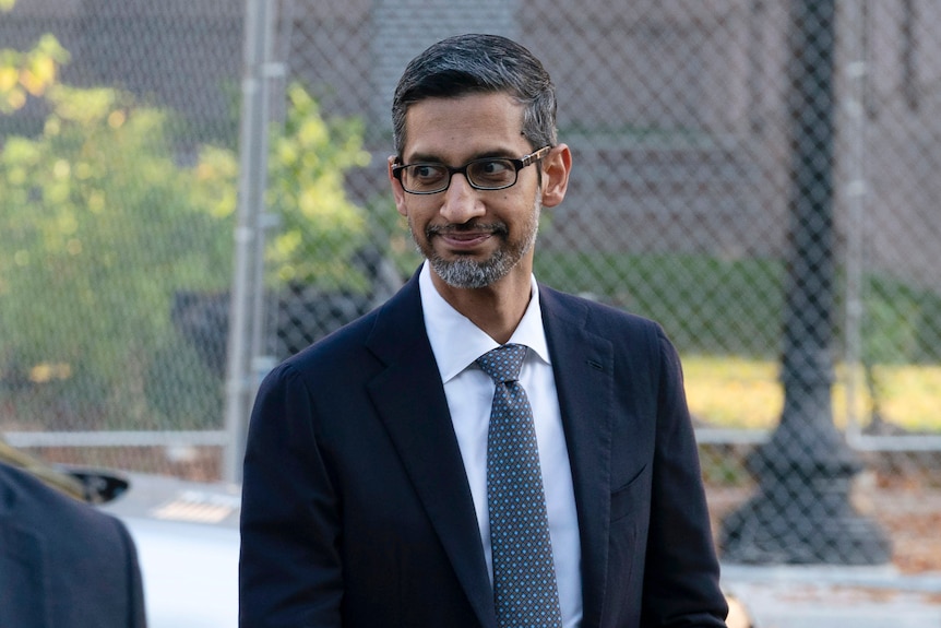 An Indian man with facial hair, in a dark suit, smiles without teeth, outside, approcahing a court.