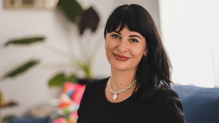 A woman with dark hair stares straight into the camera. She is sitting in her living room with a blurred out plant behind her.