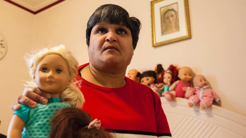 A middle-aged Indian woman in a bedroom, holding a doll, but with a sad look on her face.