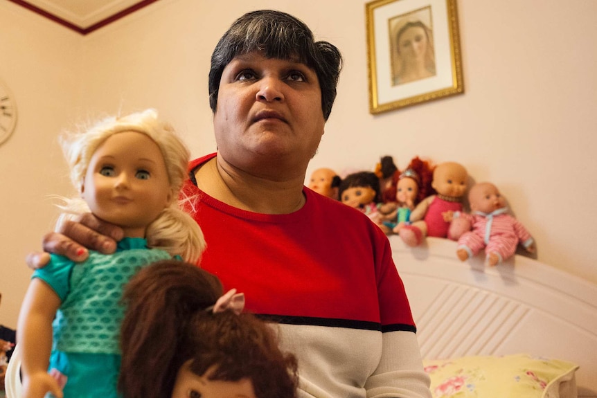 A middle-aged Indian woman in a bedroom, holding a doll, but with a sad look on her face.