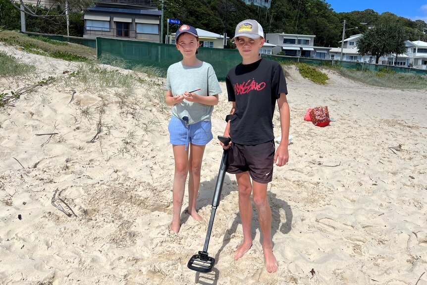 A young girl and boy wear shorts and tshirts whilst using a metal detector on a beach