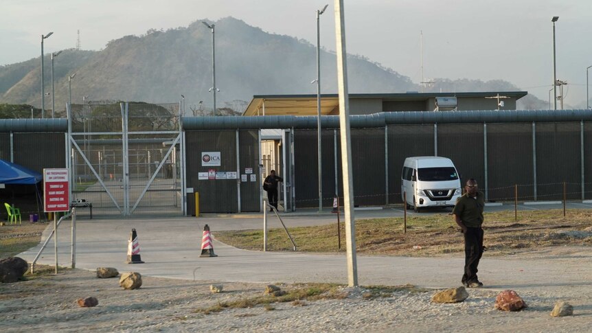 A man stands at the front of the Bomana Detention Centre. It has a high fence, a secure metal gate and light towers throughout.