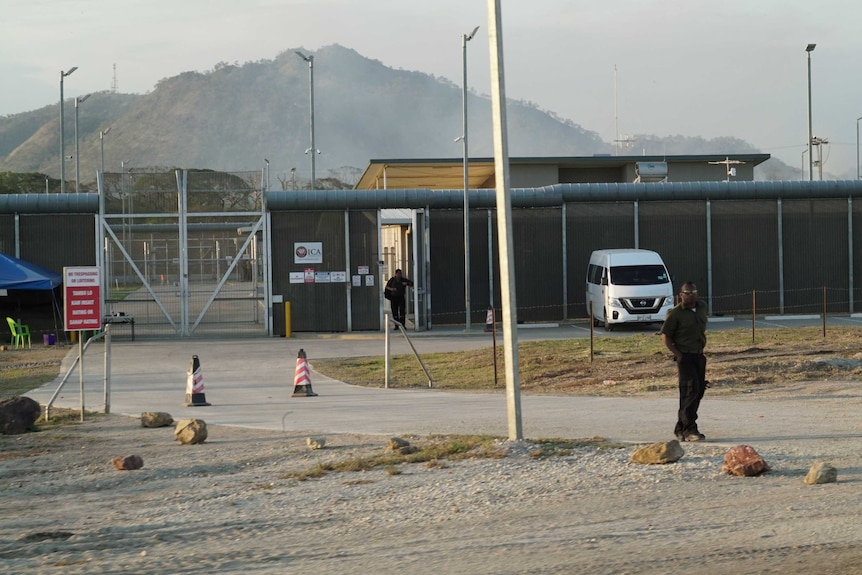A man stands at the front of the Bomana Detention Centre. It has a high fence, a secure metal gate and light towers throughout.