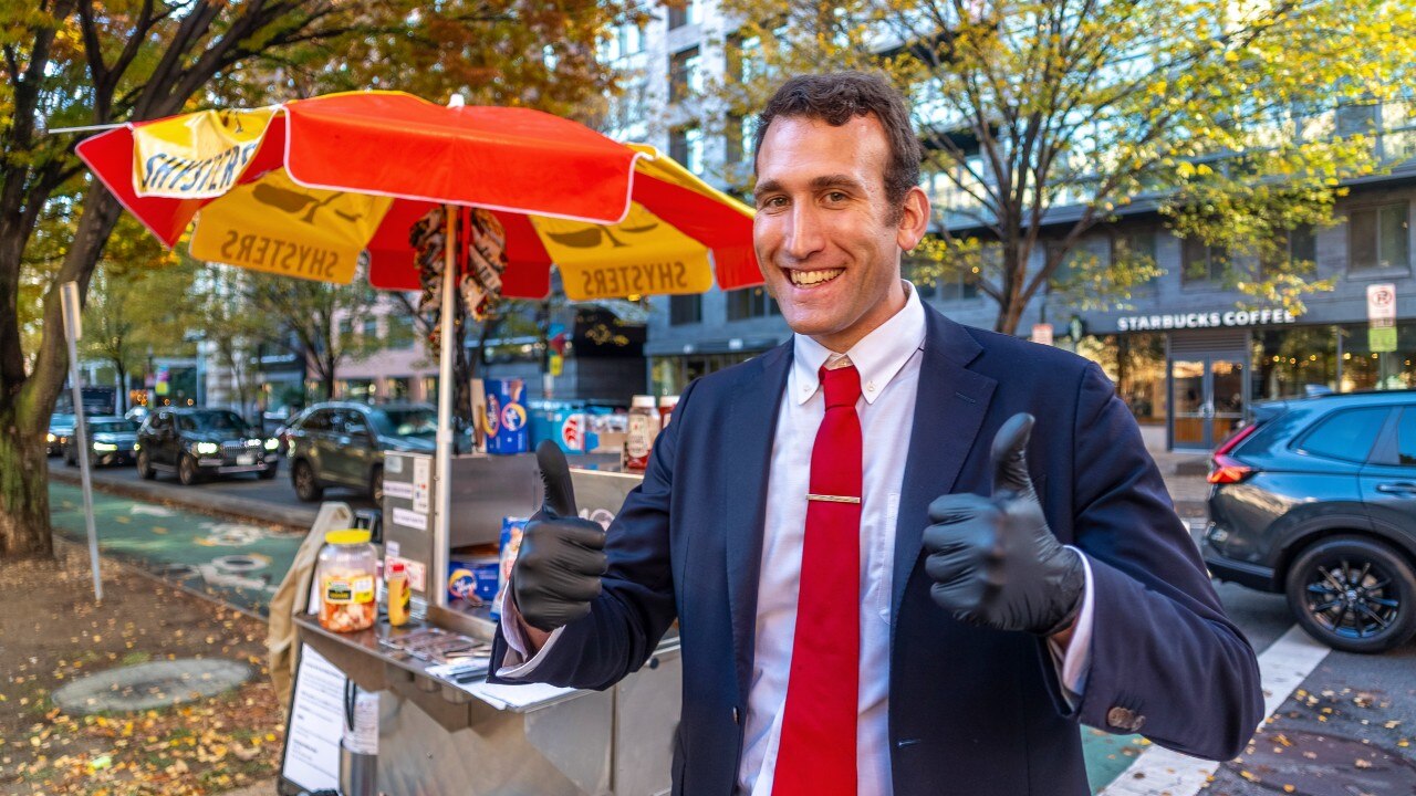 A man in a suit looking at the camera and giving the thumbs up sign with both hands. A small cart is visible in the background.