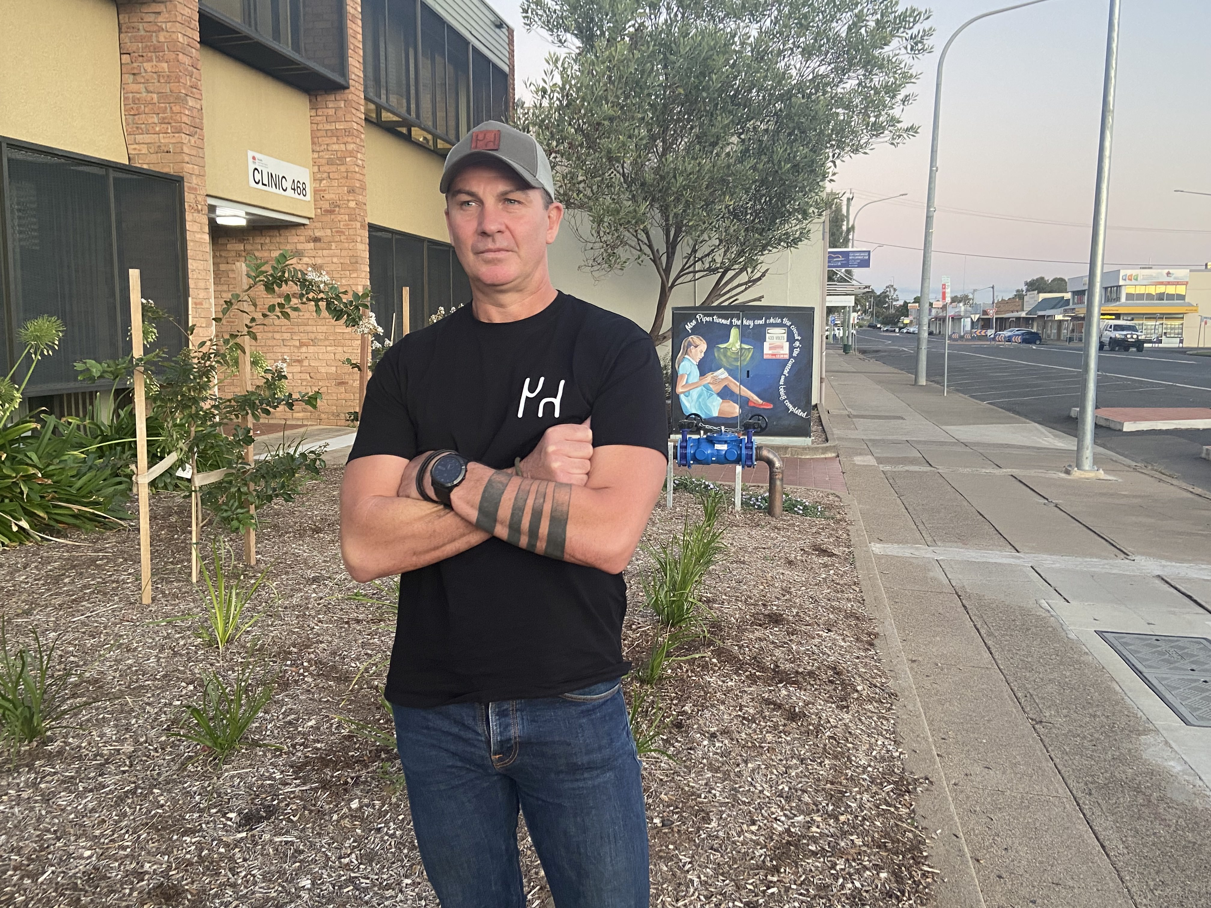 A middle aged man stands with his arms folded in front of a brick building