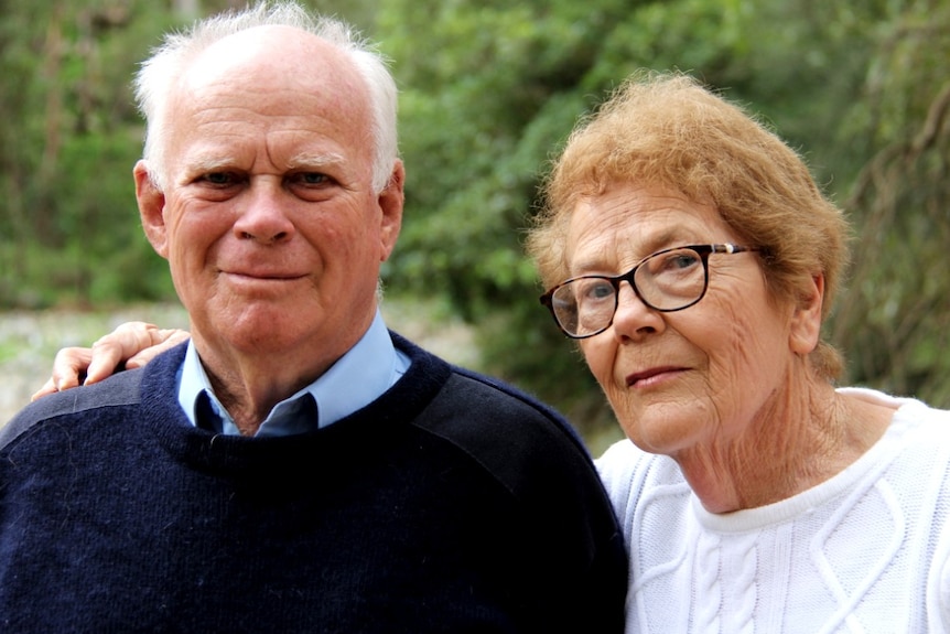 An elderly couple stand in a clearing of a rural area.