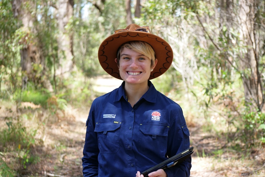 Blonde woman in the bush wearing blue NSW Government overalls with a hat on her head.