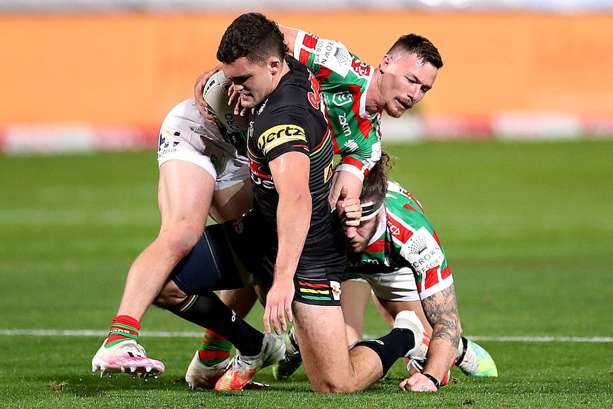 A Penrith NRL player kneels on his left knee as he is tackled by a South Sydney opponent.