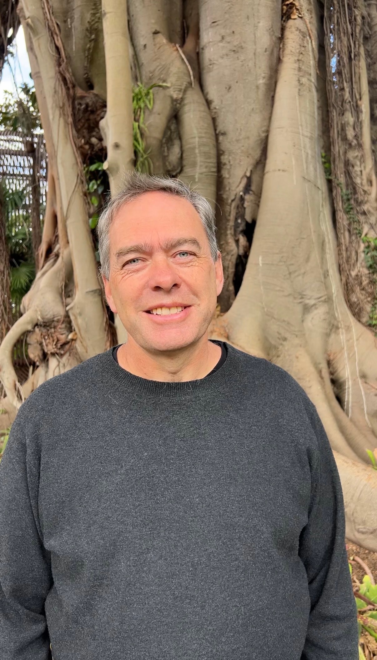A man with grey hear stands in front of a moreton bay fig trunk and smiles at the camera