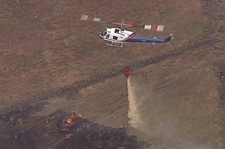 A helicopter drops a huge bucket of water over a largely burnt-out patch of grassland, with some orange flames still visible.