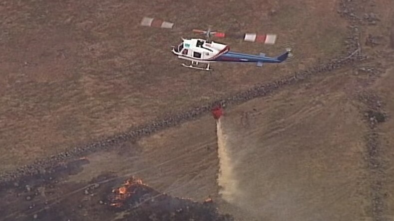 A helicopter drops a huge bucket of water over a largely burnt-out patch of grassland, with some orange flames still visible.