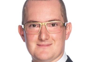 A tightly framed headshot of a man wearing golden framed glasses, a white shirt, a charcoal suit jacked and a blue tie