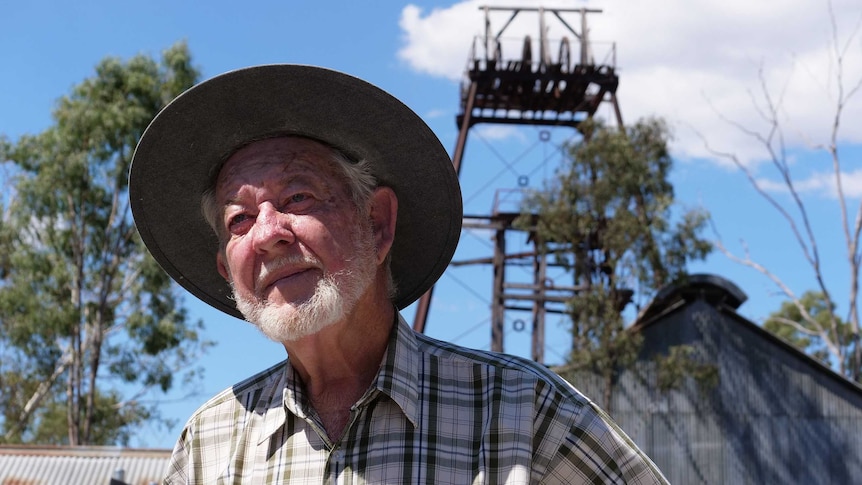 An older man stands in front of a piece of mining equipment