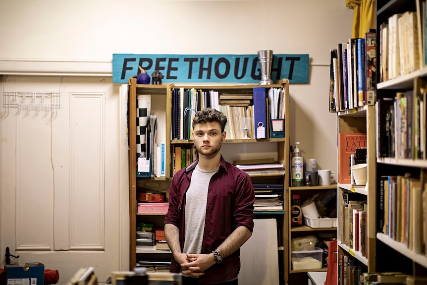 Benjamin Lynch stands in front of a cluttered bookcase with a sign on top reading "free thought."