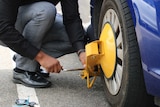 A man attaches a wheel clamp to a blue car.
