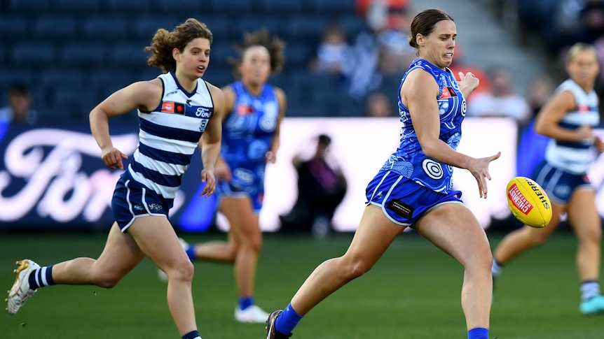 A Kangaroos AFLW player runs clear in the middle of the ground as she drops the ball to kick it as a defender trails. 