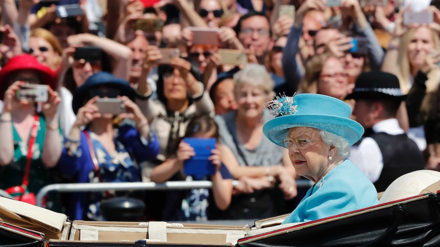 A well dressed elderly woman dressed in a blue outfit sits in an open carriage with onlookers blurred in the background