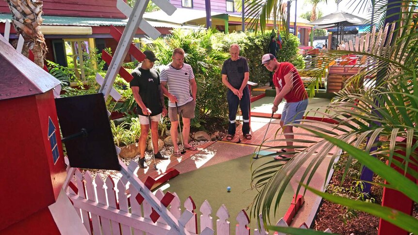 Three men stand on a mini-golf course as another man plays.