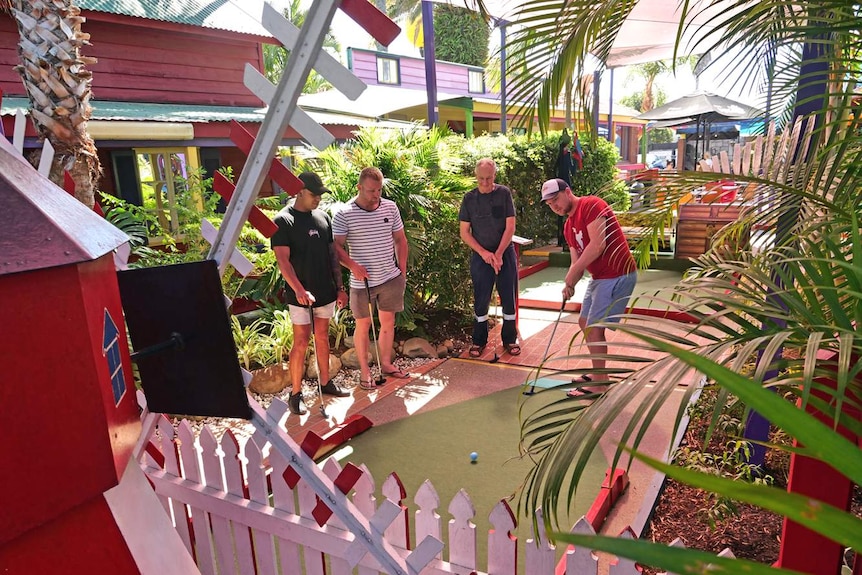 Three men stand on a mini-golf course as another man plays.