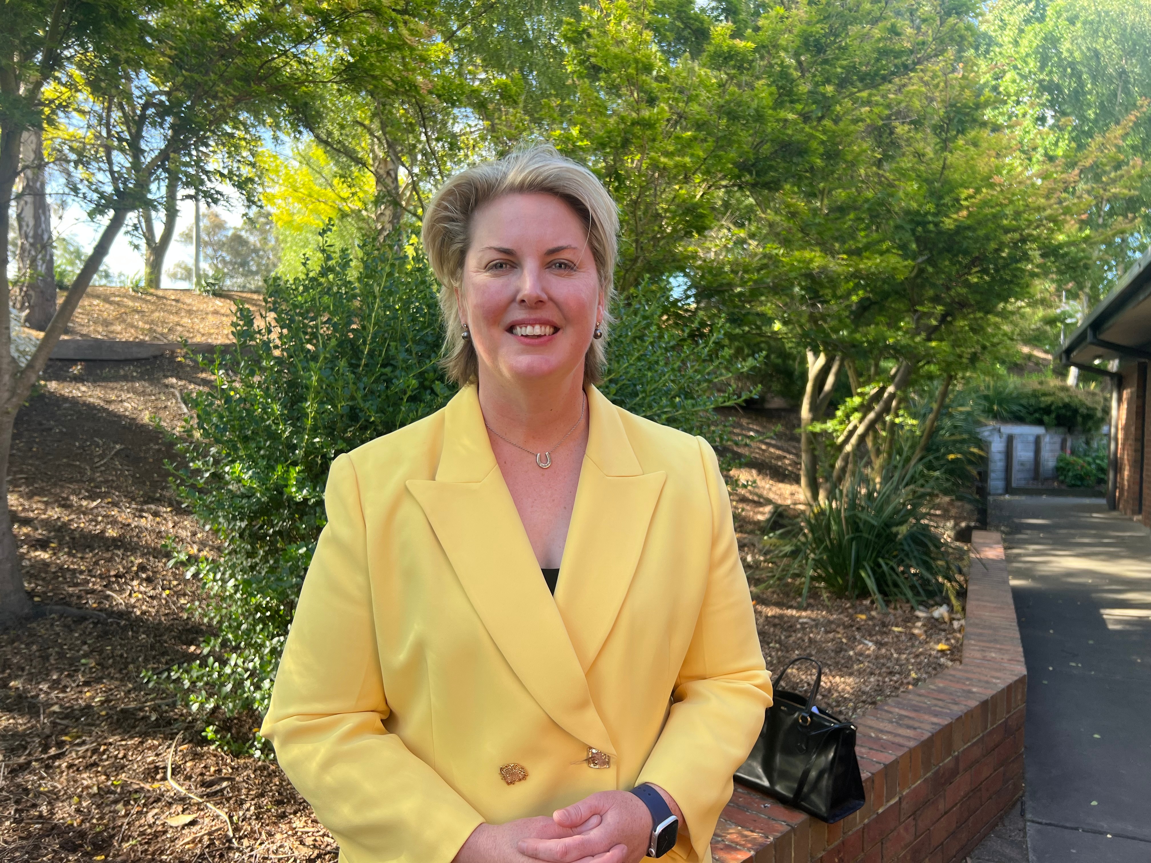 A smiling woman with short, blonde hair stands outside. She wears a brightly-coloured suit.