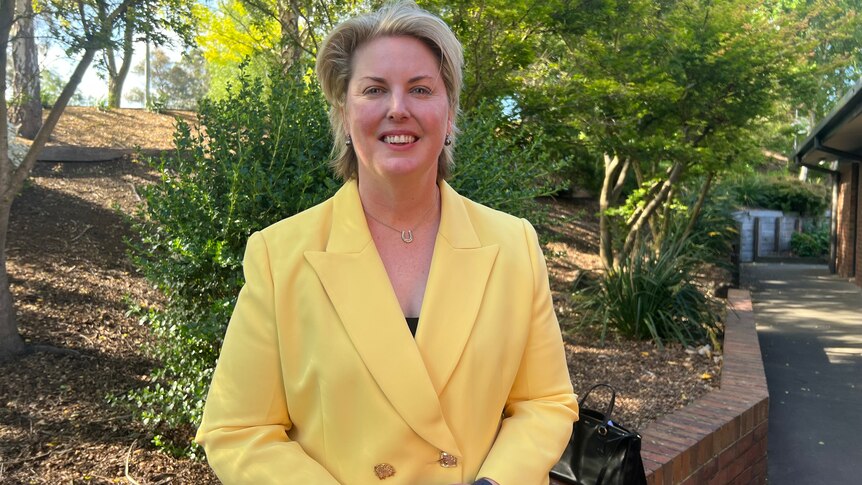 A smiling woman with short, blonde hair stands outside. She wears a brightly-coloured suit.