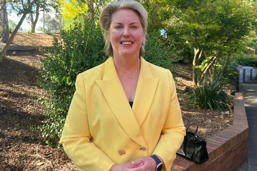 A smiling woman with short, blonde hair stands outside. She wears a brightly-coloured suit.