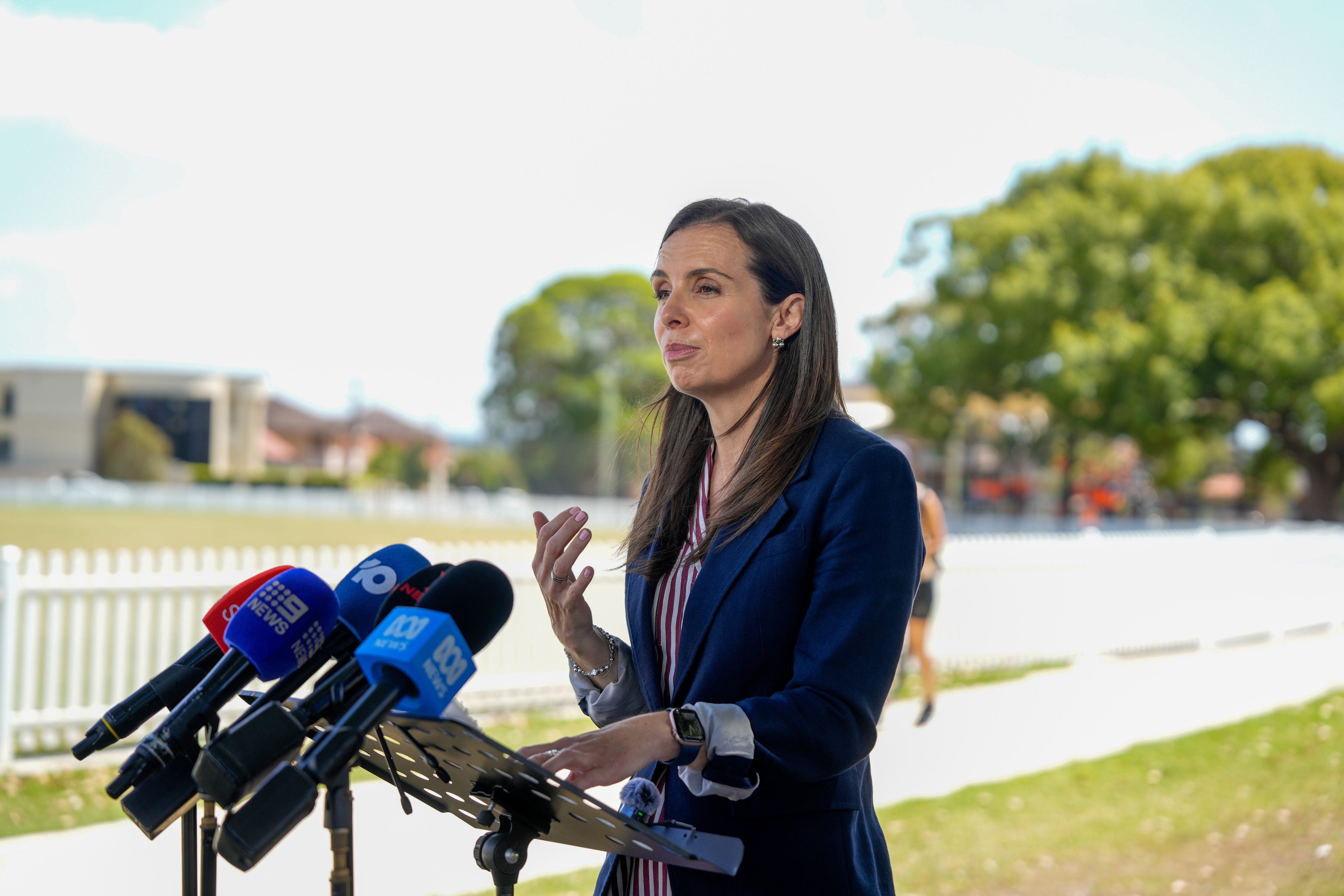 La señora Houssos habla ante los micrófonos de los medios de comunicación en una conferencia de prensa junto a un óvalo de cricket.