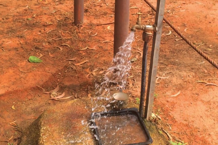 A rusty old outside tap in red dirt turned on and gushing water.